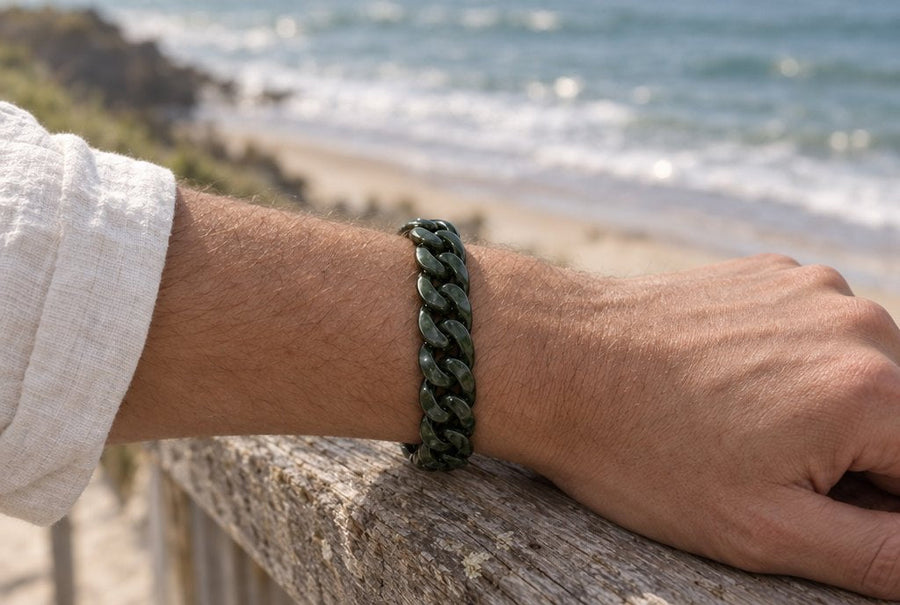 Forearm wearing a dark green jade Cuban bracelet resting on a timber railing above a beach